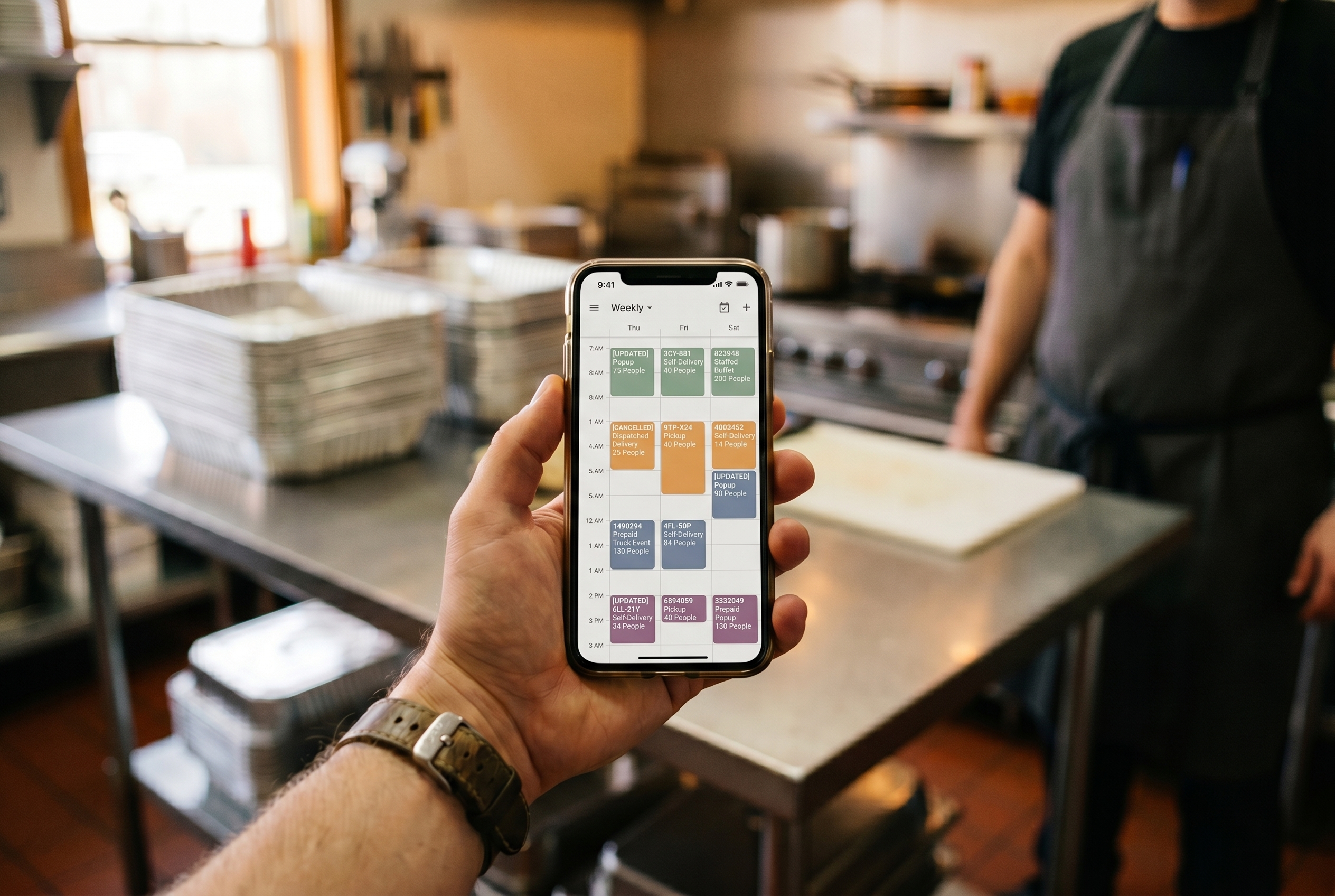 A hand holding a phone showing a color-coded weekly catering calendar